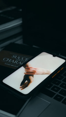 A smartphone displaying an image of two hands touching is placed on a laptop keyboard. In the background, a book with partially visible text is also present.