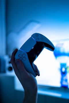 A hand is holding a modern gaming controller, illuminated by a blue-tinted light. The background is blurred, possibly showing a gaming console or television.