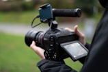A person holding a professional video camera, focusing on a touchscreen display. The camera features a long lens and a handle, suggesting it is used for filming high-quality videos. The background is blurred, indicating the use of depth of field.