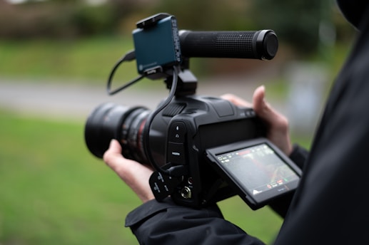 A person holding a professional video camera, focusing on a touchscreen display. The camera features a long lens and a handle, suggesting it is used for filming high-quality videos. The background is blurred, indicating the use of depth of field.