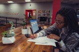 A person with braided hair and glasses is sitting at a wooden table, writing on some papers. There is an open laptop beside them, along with two potted plants on the table. The background shows a room with artwork on the walls and another person walking past an open doorway.