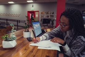 A person with braided hair and glasses is sitting at a wooden table, writing on some papers. There is an open laptop beside them, along with two potted plants on the table. The background shows a room with artwork on the walls and another person walking past an open doorway.