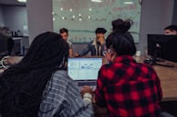 A group of people are gathered around a table with computers in a coworking space. Two individuals in the foreground are focused on a laptop displaying spreadsheets or data. A glass wall in the background is decorated with drawings and the text 'Base Camp Coworking', adding to the collaborative environment.