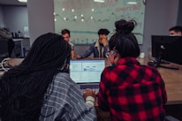 A group of people are gathered around a table with computers in a coworking space. Two individuals in the foreground are focused on a laptop displaying spreadsheets or data. A glass wall in the background is decorated with drawings and the text 'Base Camp Coworking', adding to the collaborative environment.