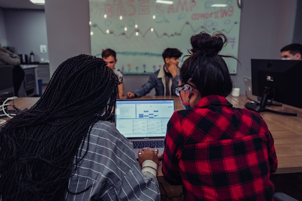 A group of people are gathered around a table with computers in a coworking space. Two individuals in the foreground are focused on a laptop displaying spreadsheets or data. A glass wall in the background is decorated with drawings and the text 'Base Camp Coworking', adding to the collaborative environment.