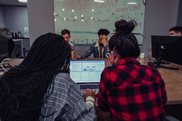 A group of people are gathered around a table with computers in a coworking space. Two individuals in the foreground are focused on a laptop displaying spreadsheets or data. A glass wall in the background is decorated with drawings and the text 'Base Camp Coworking', adding to the collaborative environment.