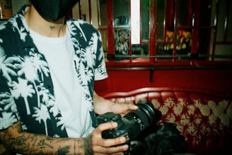 A person holding a DSLR camera with both hands is partially visible, wearing a palm tree-patterned shirt and a white undershirt. Their left arm has visible tattoos, and they are seated on a red patterned couch. A mirror reflects parts of the scene in the background.