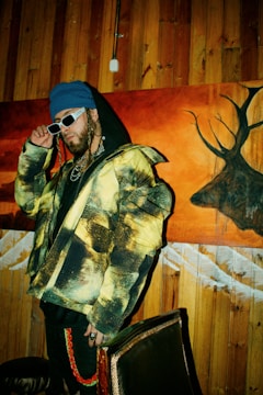 A person wearing a colorful jacket with a yellow and black pattern, a blue beanie, and white sunglasses stands in front of a wooden wall and a vibrant painting featuring a deer silhouette. They exude a stylish, urban vibe with braided hair and multiple chains around their neck.