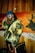 A person wearing a colorful jacket with a yellow and black pattern, a blue beanie, and white sunglasses stands in front of a wooden wall and a vibrant painting featuring a deer silhouette. They exude a stylish, urban vibe with braided hair and multiple chains around their neck.