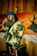 A person wearing a colorful jacket with a yellow and black pattern, a blue beanie, and white sunglasses stands in front of a wooden wall and a vibrant painting featuring a deer silhouette. They exude a stylish, urban vibe with braided hair and multiple chains around their neck.