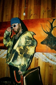 A person wearing a colorful jacket with a yellow and black pattern, a blue beanie, and white sunglasses stands in front of a wooden wall and a vibrant painting featuring a deer silhouette. They exude a stylish, urban vibe with braided hair and multiple chains around their neck.