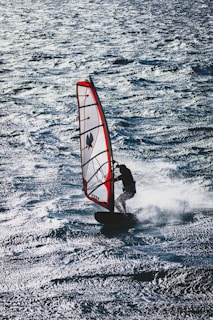 A windsurfer skillfully navigates the choppy waters, balancing on a board with a red and white sail. The sunlight reflects off the waves, creating a shimmering effect on the surface of the sea.