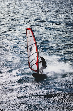 A windsurfer skillfully navigates the choppy waters, balancing on a board with a red and white sail. The sunlight reflects off the waves, creating a shimmering effect on the surface of the sea.