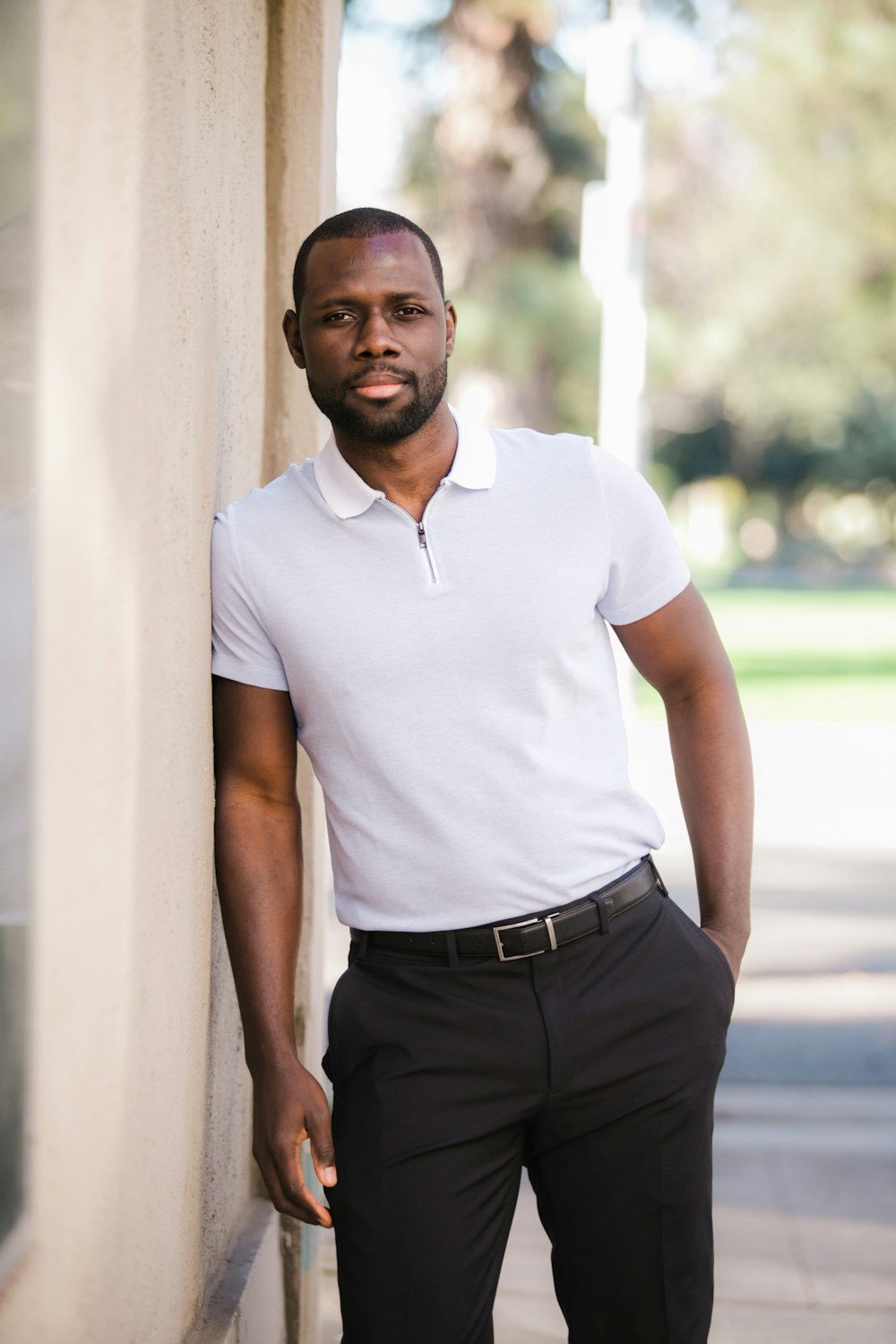 Man in white polo shirt and black pants standing beside white wall