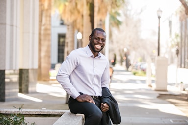 photography poses for men,how to photograph fortune vieyra sitting and smiling with black vest in hand.; man in white dress shirt and black pants sitting on the sidewalk during daytime