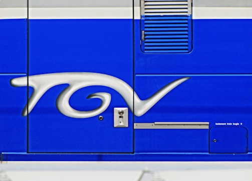 A close-up of a blue surface featuring a stylized silver logo or emblem with flowing curves. The background includes mechanical elements like a vent and a small access panel.