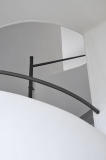 A minimalist architectural space with white walls and a simple, black metal railing curving along the edge of a staircase. The lighting creates soft shadows, enhancing the smooth lines and subtle curves of the design.
