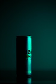 A can with a gradient of green lighting casts dramatic shadows in a dimly lit setting. The can appears to be a product shot with reflections and condensation visible, suggesting a cold beverage.