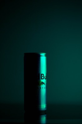 A can with a gradient of green lighting casts dramatic shadows in a dimly lit setting. The can appears to be a product shot with reflections and condensation visible, suggesting a cold beverage.