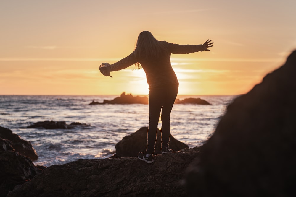 silhouette of woman standing on rock near sea during sunset