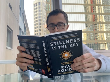 A person with glasses is seated at a glass table, reading a book titled 'Stillness is the Key' by Ryan Holiday. The background features tall buildings with many windows, suggesting an urban setting. The person appears focused on the book, which has a black cover with white and yellow text and graphics.