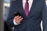 A person wearing a dark blue business suit with a white shirt and a purple tie is holding a smartphone in their left hand. The background is blurred, suggesting an indoor setting.