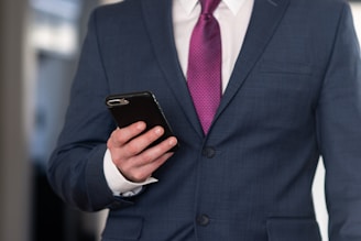 A person wearing a dark blue business suit with a white shirt and a purple tie is holding a smartphone in their left hand. The background is blurred, suggesting an indoor setting.