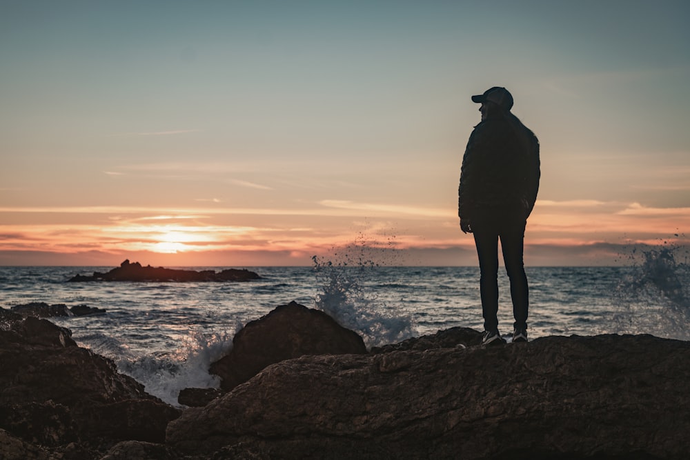 silhouette of man standing on rock formation near body of water during sunset