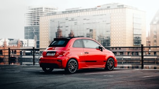 A small, red sports car is parked in an outdoor lot with a cityscape in the background. The car has black wheels, tinted windows, and distinct branding on the side. The car is situated near a railing overlooking a reflective water body, with modern and historic buildings visible in the distance.