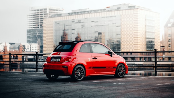 A small, red sports car is parked in an outdoor lot with a cityscape in the background. The car has black wheels, tinted windows, and distinct branding on the side. The car is situated near a railing overlooking a reflective water body, with modern and historic buildings visible in the distance.
