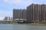 A waterfront cityscape featuring numerous tall, densely packed residential high-rise buildings. The structures are arranged closely together, showcasing a modern urban design with various balconies and windows. In the foreground, a body of water reflects the skyline, bordered by a few trees and a small pavilion along the shoreline.