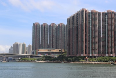 A waterfront cityscape featuring numerous tall, densely packed residential high-rise buildings. The structures are arranged closely together, showcasing a modern urban design with various balconies and windows. In the foreground, a body of water reflects the skyline, bordered by a few trees and a small pavilion along the shoreline.