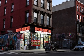 A corner deli with a brightly lit storefront displays various snacks and drinks inside. The exterior features vibrant street art and graffiti covering the adjacent brick walls. A few bicycles are parked nearby, and an evening urban environment is evident from the dim street lighting.