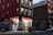 A corner deli with a brightly lit storefront displays various snacks and drinks inside. The exterior features vibrant street art and graffiti covering the adjacent brick walls. A few bicycles are parked nearby, and an evening urban environment is evident from the dim street lighting.