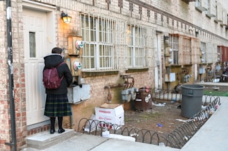 A person wearing a dark jacket and a plaid skirt stands on a small step in front of a residential door. They have a maroon backpack and appear to be interacting with a utility meter next to the door. The building is made of brick with barred windows. Below, on the ground, there are small metal fences, brown trash bins, and some scattered debris. A cardboard box labeled 'Red Chief' is also visible.