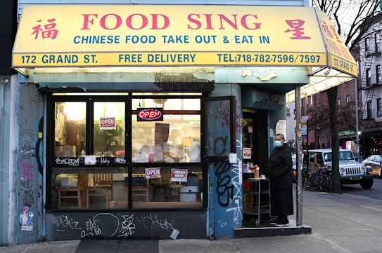 A Chinese food takeout and dine-in restaurant named 'Food Sing' located at 172 Grand St. The storefront features a large yellow canopy with text in both English and Chinese. The entrance has a glass door and windows where several 'OPEN' signs are displayed. There is noticeable graffiti on the lower portion of the establishment's facade. A person wearing a face mask stands near the entrance.
