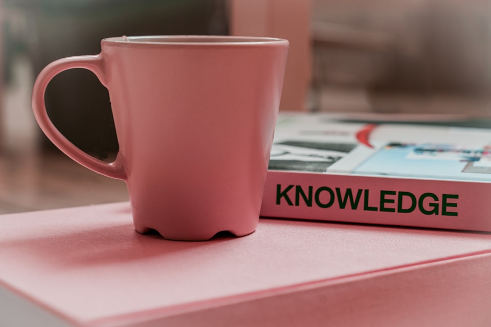 Close-up of a pink coffee mug next to a book with the word "knowledge" resting on a table.