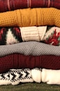A stack of neatly folded knitted sweaters with various textures and patterns, showing a range of colors from deep red and mustard yellow to gray, burgundy, and patterned white and black. The fibers and knitting patterns are clearly visible, emphasizing the cozy and warm nature of the garments.