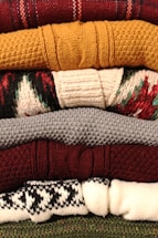 A stack of neatly folded knitted sweaters with various textures and patterns, showing a range of colors from deep red and mustard yellow to gray, burgundy, and patterned white and black. The fibers and knitting patterns are clearly visible, emphasizing the cozy and warm nature of the garments.