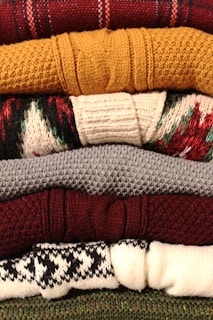 A stack of neatly folded knitted sweaters with various textures and patterns, showing a range of colors from deep red and mustard yellow to gray, burgundy, and patterned white and black. The fibers and knitting patterns are clearly visible, emphasizing the cozy and warm nature of the garments.