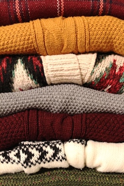 A stack of neatly folded knitted sweaters with various textures and patterns, showing a range of colors from deep red and mustard yellow to gray, burgundy, and patterned white and black. The fibers and knitting patterns are clearly visible, emphasizing the cozy and warm nature of the garments.