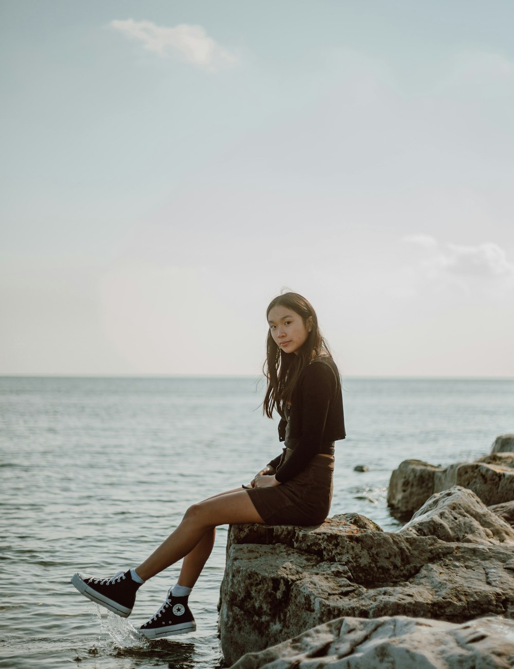 woman in black long sleeve dress sitting on rock near sea during daytime