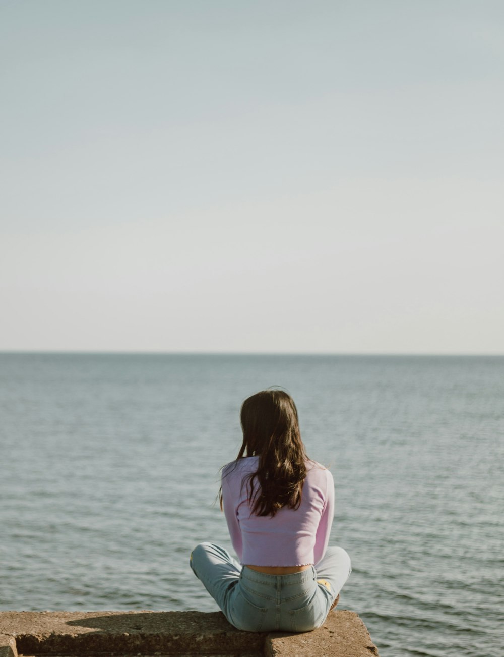 woman in pink long sleeve shirt sitting on gray rock near body of water during daytime
