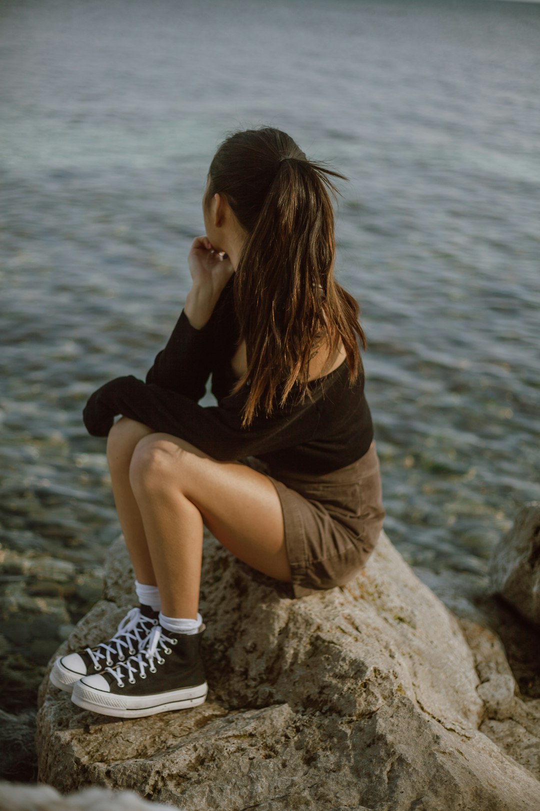 woman in black long sleeve shirt and white shorts sitting on rock near body of water