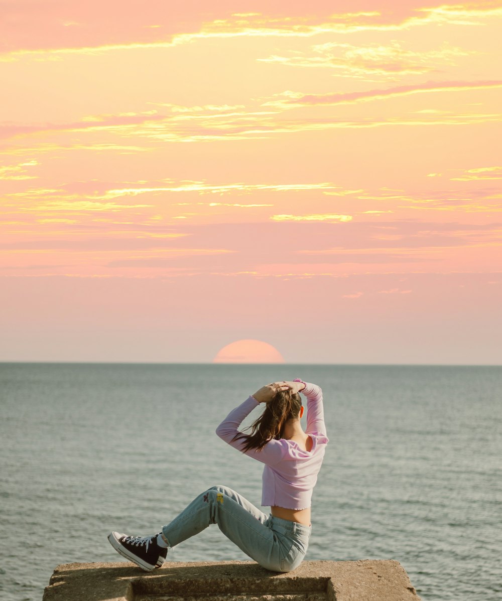 woman in white dress sitting on gray rock near body of water during sunset