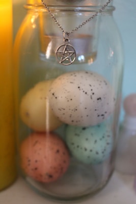 A glass jar contains four speckled eggs in pastel colors: yellow, white, turquoise, and pink. A silver pentagram pendant is hanging at the top of the jar. In the background, there is a yellow candle and a small bottle.