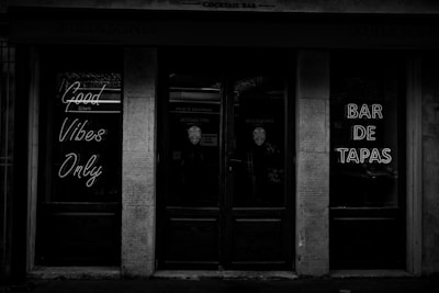 A black-and-white photo of a storefront entrance with neon signs. The signs read 'Good Vibes Only' and 'Bar de Tapas'. The atmosphere appears dark and moody, created by minimal lighting and reflective glass surfaces.