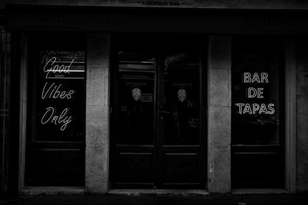 A black-and-white photo of a storefront entrance with neon signs. The signs read 'Good Vibes Only' and 'Bar de Tapas'. The atmosphere appears dark and moody, created by minimal lighting and reflective glass surfaces.