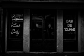 A black-and-white photo of a storefront entrance with neon signs. The signs read 'Good Vibes Only' and 'Bar de Tapas'. The atmosphere appears dark and moody, created by minimal lighting and reflective glass surfaces.
