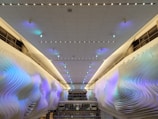 A modern interior with an illuminated ceiling covered in small, recessed lights. The side walls display an artistic, wave-like texture with soft, pastel blue and purple hues. Large windows can be seen at the far end, partially reflecting the ambient light.